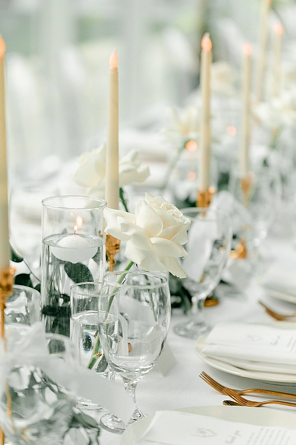 Reception tablescape with white and gold tablescape details, white roses and taper candles in glass vases over greenery garland on white linens
