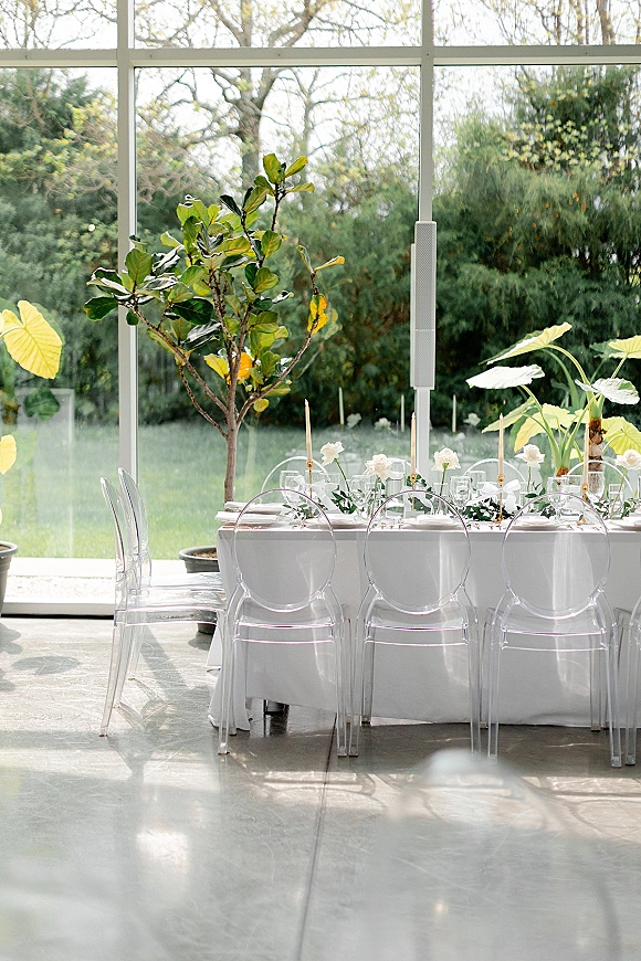 Reception tablescape with a white wedding tablescape, clear acrylic chairs, greenery garland and taper candles by floor-to-ceiling windows overlooking a garden view