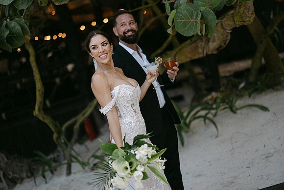 Couple portrait of bride and groom with cocktails, her lace strapless gown and bouquet, under string lights amid leafy branches on sand