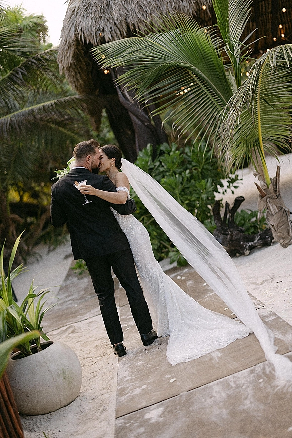 Wedding kiss portrait of bride and groom kiss, her long veil and lace dress flowing as they toast with martinis beneath palm trees