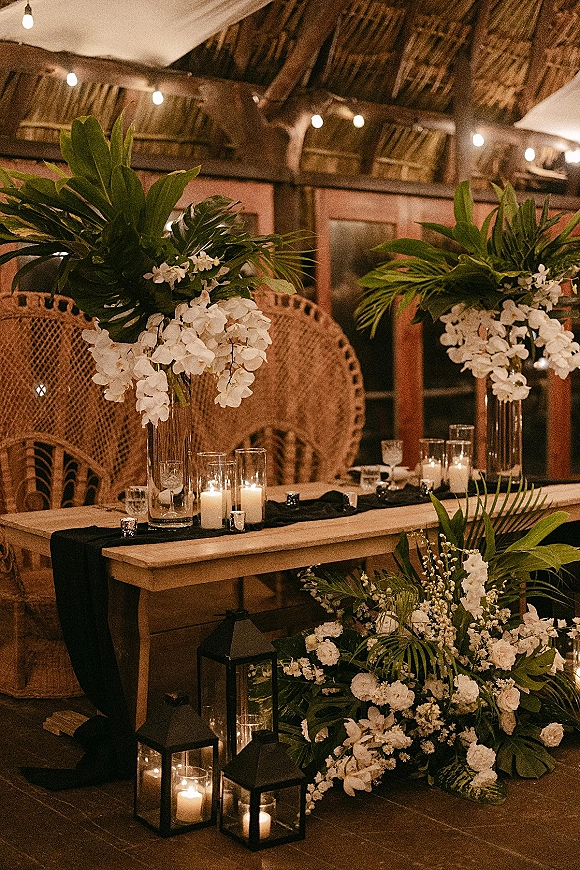 Sweetheart table decor with white florals and tropical greenery in tall glass vases, candlelit on a wooden table beneath string lights and a thatched ceiling