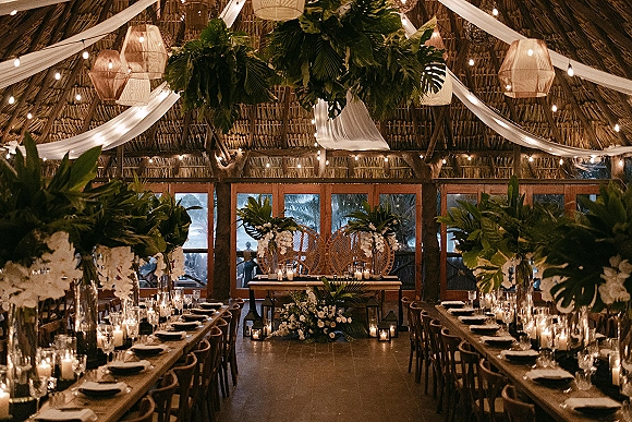 Reception tablescape with a long table wedding reception setup, candles and lanterns, tropical greenery, and string lights under a thatched ceiling