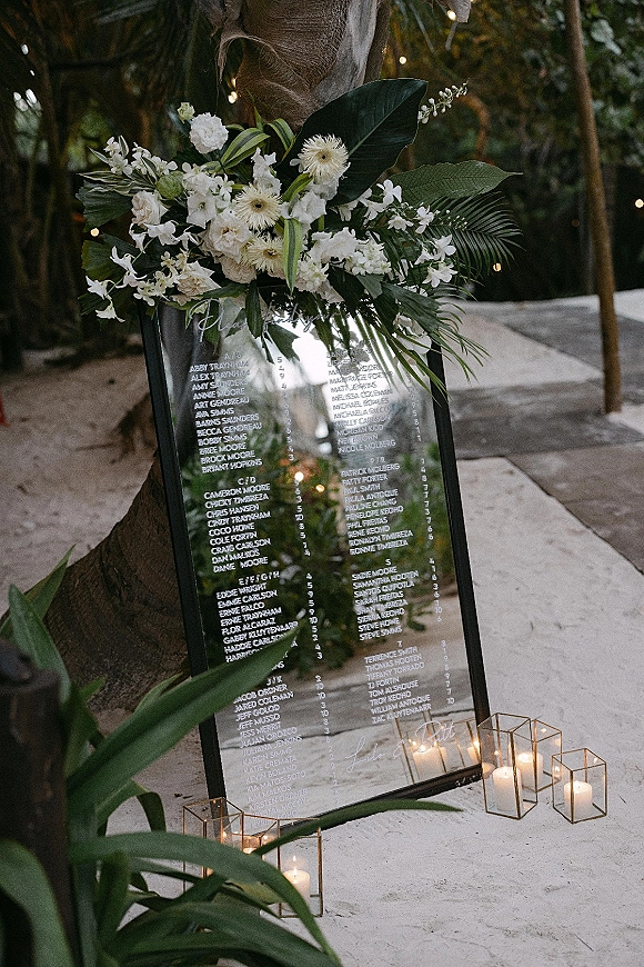 Wedding seating chart mirror sign with calligraphy, white flowers, and candles on sand with palm trees and string lights behind