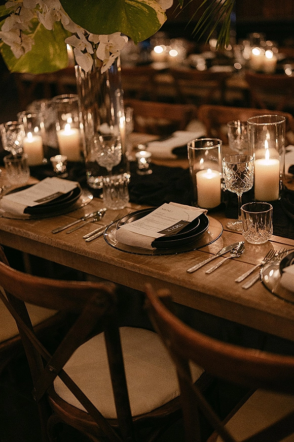 Reception tablescape with a candlelit reception table on a wood farm table, black plates, silver flatware, greenery centerpiece in dim light