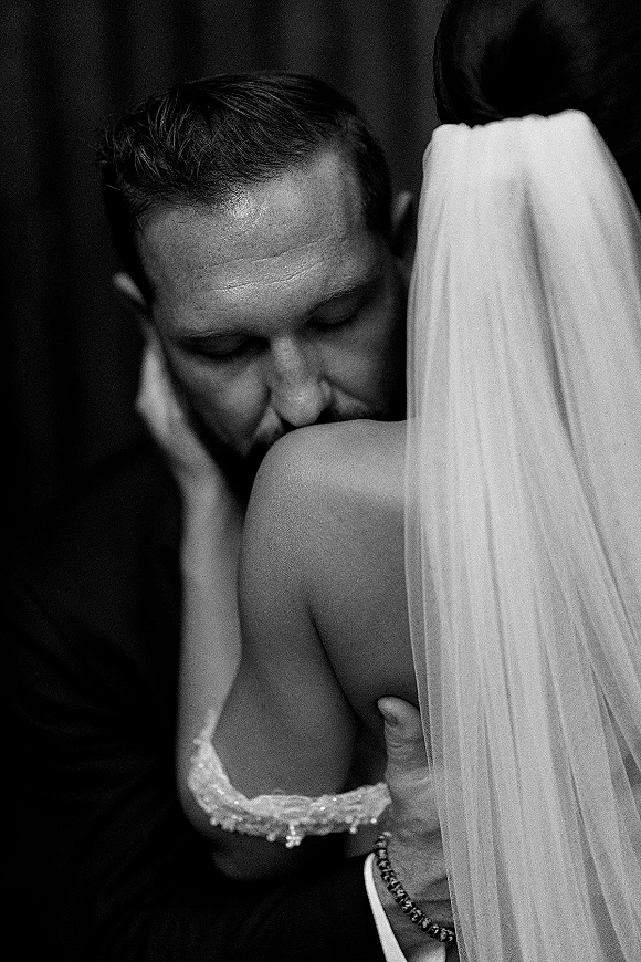 Wedding couple portrait in a black and white wedding photo, groom kissing the bride’s shoulder as she hugs him, veil draped against a dark blurred backdrop