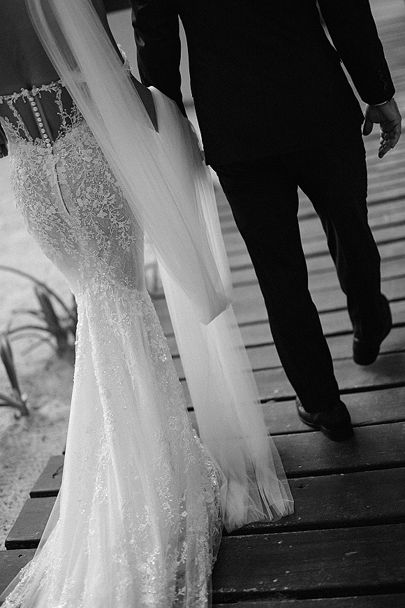 Couple walking away with wedding veil train flowing behind a lace mermaid wedding dress on a wooden boardwalk through sand and plants