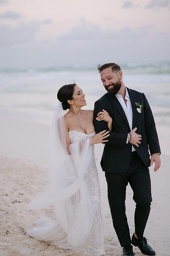Couple portrait of bride and groom at a beach wedding couple stroll, her lace gown and veil blowing in ocean breeze by waves