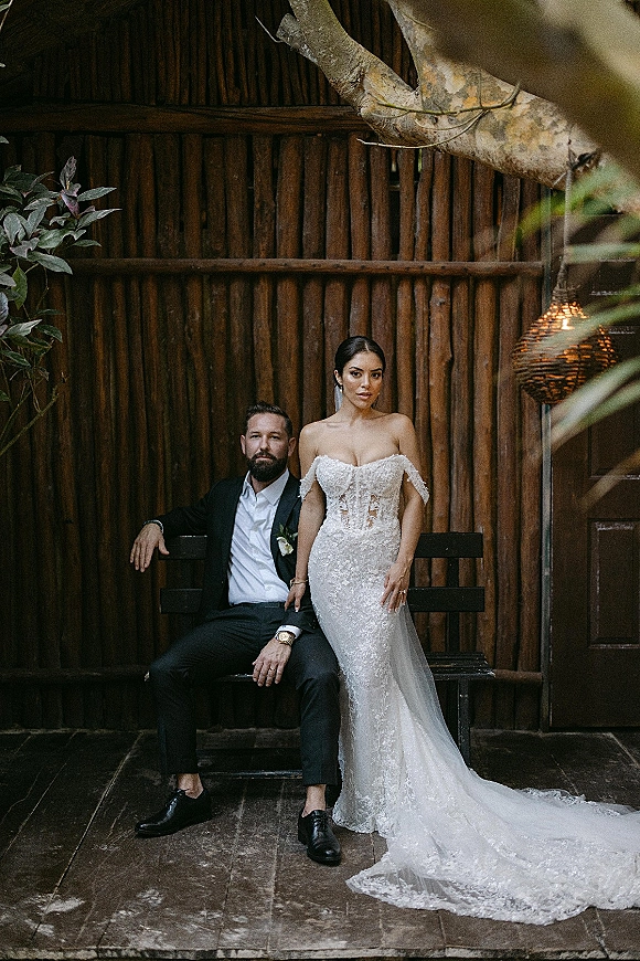 Couple portrait of bride in off-the-shoulder lace dress and veil standing by groom in black suit on a wooden bench, tropical wood wall backdrop