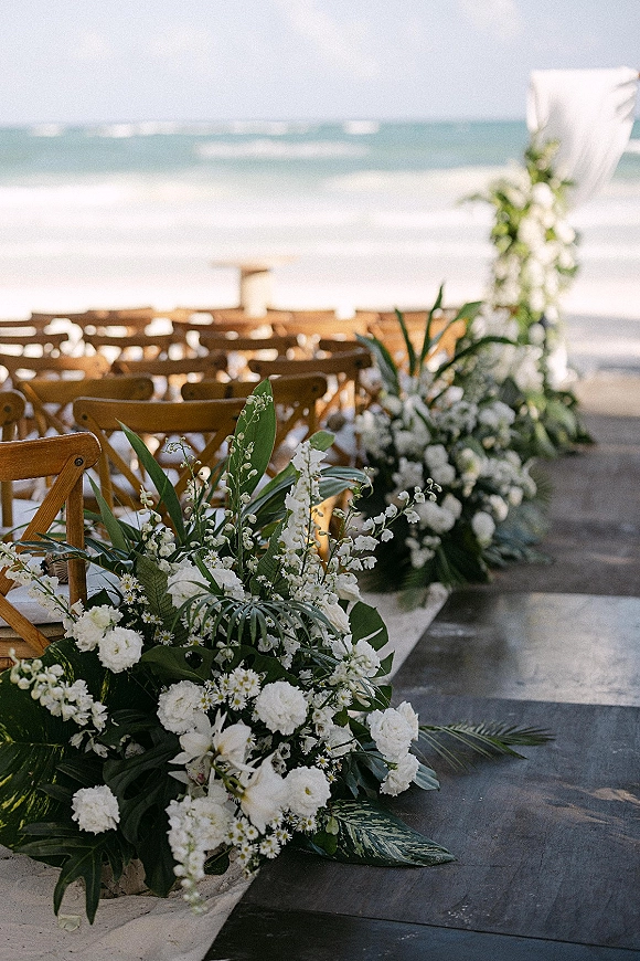 Beach ceremony setup with an aisle runner, white floral arrangements and tropical greenery leading to a draped arch by the ocean