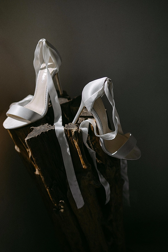 Bridal shoes with white satin ribbon ankle ties rest on a wood stump, lit by window light against a dark wall with shadows
