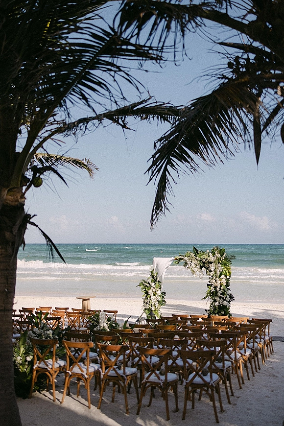 Beach ceremony setup with an oceanfront wedding ceremony arch draped in white fabric and tropical greenery, chairs facing waves and palms