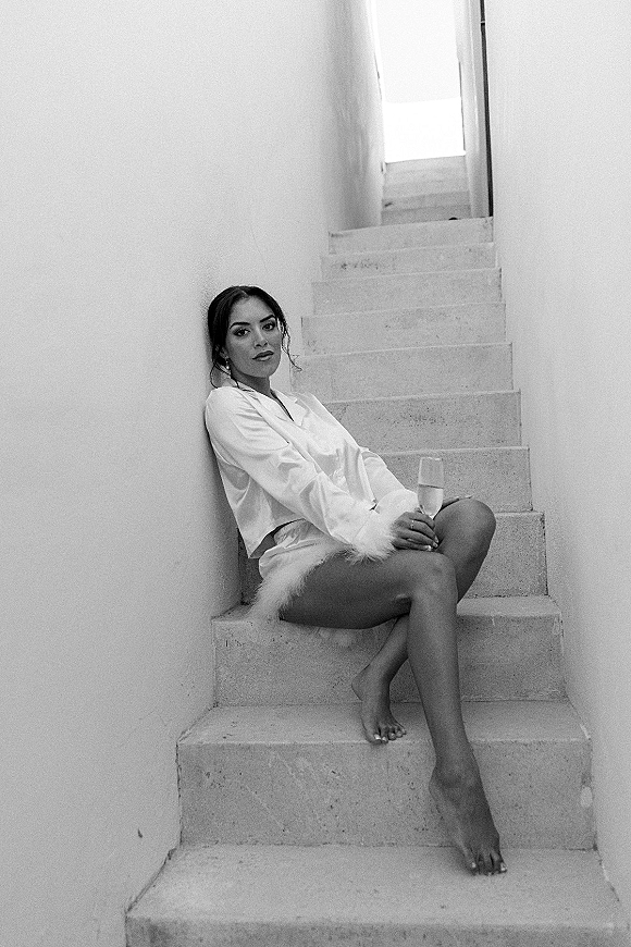 Bridal boudoir portrait of a bride in wedding getting ready pajamas with feather-trim silk set, champagne flute, on a bright staircase