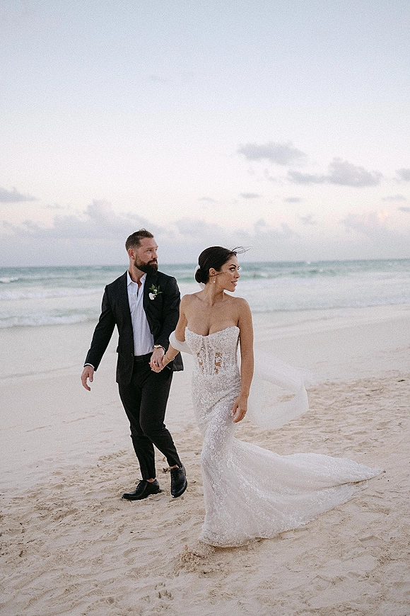 Couple portrait of a beach wedding couple holding hands on the shoreline, bride in an off-the-shoulder lace gown with train under cloudy sky