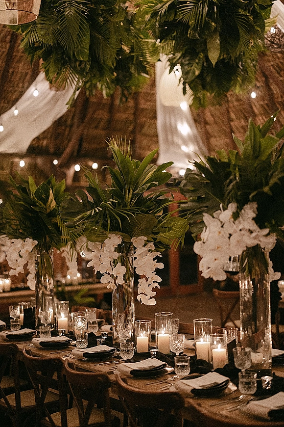 Reception tablescape with a candlelit wedding table, white orchids and tropical greenery in tall glass vases against exposed brick and string lights