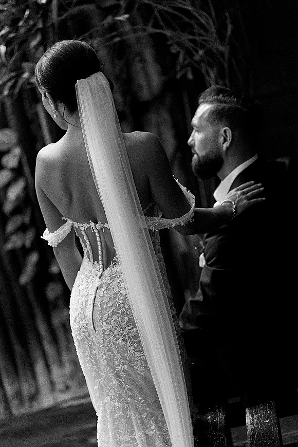 Couple portrait in a black and white wedding portrait, bride in lace off-shoulder gown and long veil embraces groom in suit by trees