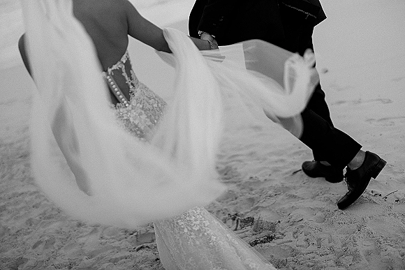 Wedding couple portrait on beach sand, groom holding the bride’s blowing veil over her lace gown as they sit by the shoreline