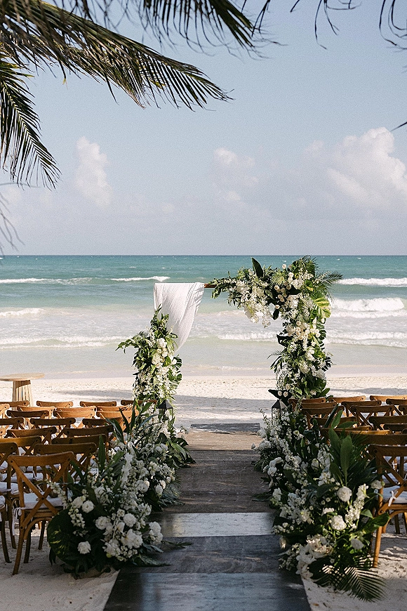 Beach ceremony setup with tropical wedding arch draped in white fabric and flowers, wooden aisle runner and chairs facing the ocean