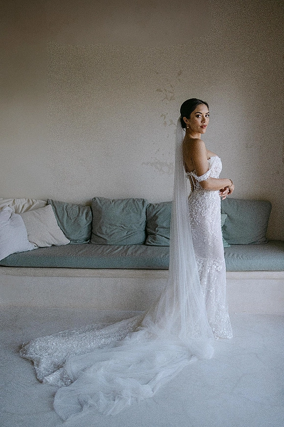 Bridal portrait of a bride looking over her shoulder in a lace wedding dress with long veil and train beside a neutral sofa indoors