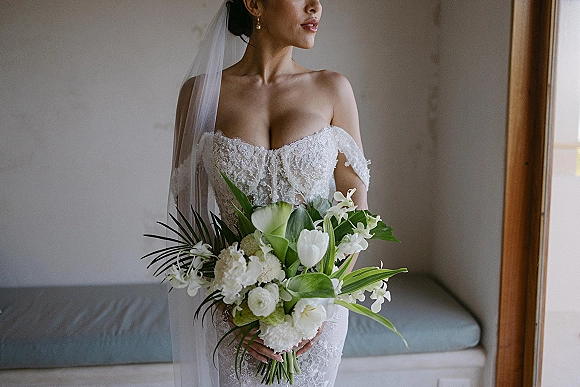 Bridal portrait of a bride in an off the shoulder wedding dress holding a bouquet, veil draped in window light by a bench seat