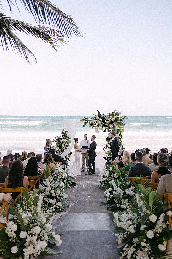 Wedding ceremony at a beach wedding ceremony with a white draped floral arch, aisle flowers, and wooden chairs facing the ocean waves