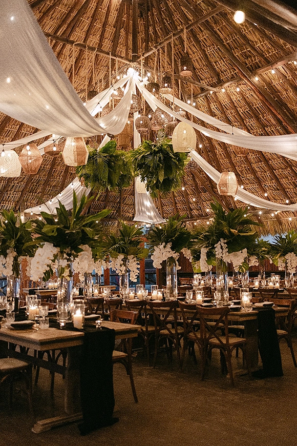 Reception tablescape with candlelit reception tables featuring white floral centerpieces and tropical greenery under string lights in a thatched pavilion