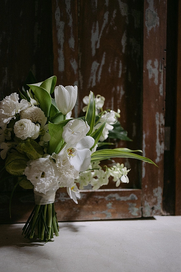 Bridal bouquet of white flowers with tulips and orchids, wrapped with greenery and resting by a rustic wooden door on a white tabletop