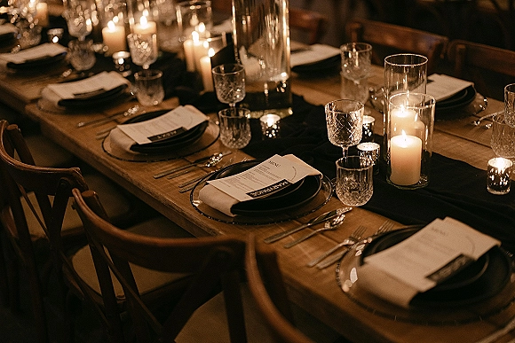 Reception tablescape with a candlelit wedding table and black table runner, pillar and votive candles glowing on a wooden banquet table in dim light