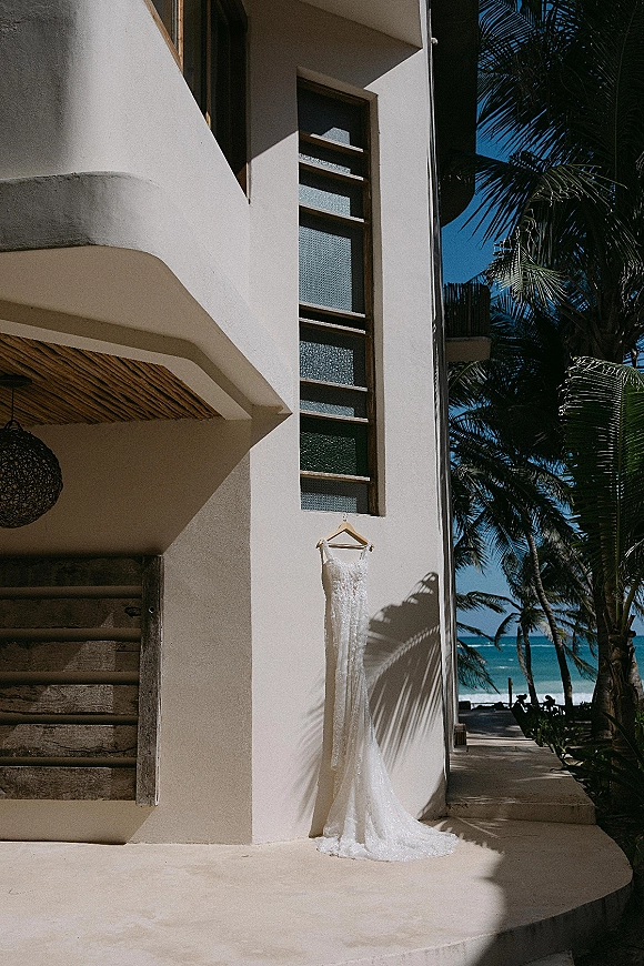 Wedding dress with beaded lace details on a wooden hanger, hanging against a white stucco wall with palms and ocean view behind
