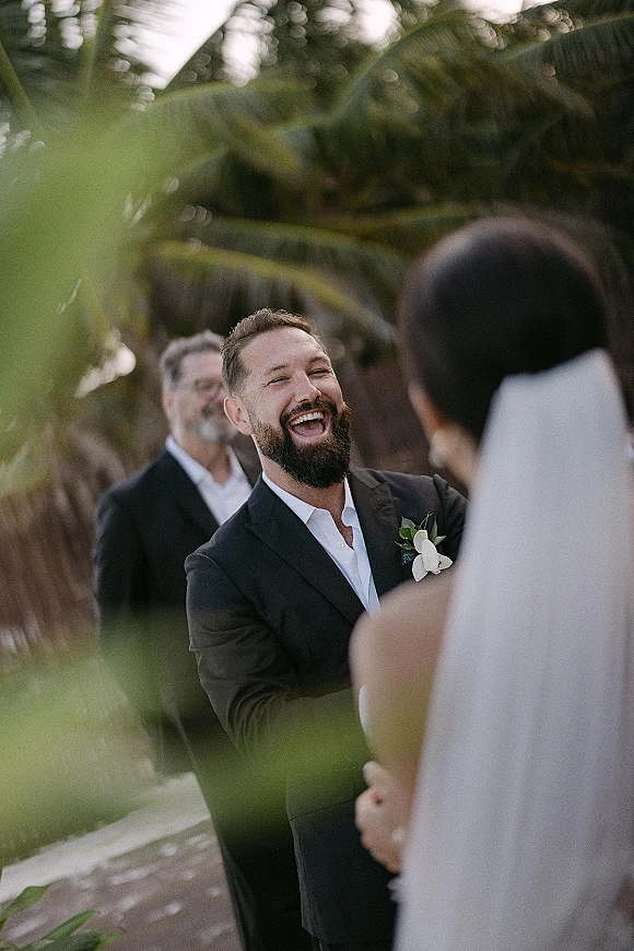 First look moment as the groom laughs while the bride approaches in her veil on a palm-lined walkway with greenery and guests behind