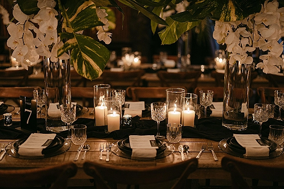 Reception tablescape with candlelit wedding table details—white orchid centerpieces in tall glass vases, black plates and napkins on wood tables