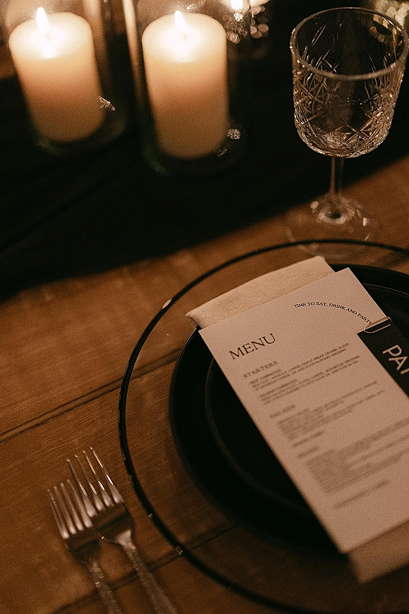 Wedding tablescape with a wedding menu card clipped to a neutral napkin on black plates, flanked by pillar candles and glass votives on a wood table