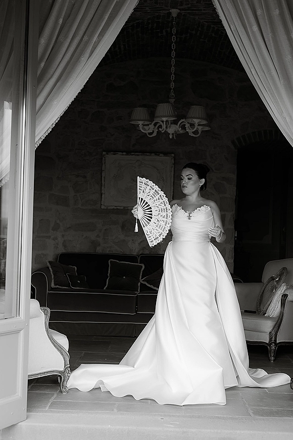 Bridal portrait of a bride holding lace fan in a strapless lace wedding dress, posed in a doorway with curtains and chandelier behind