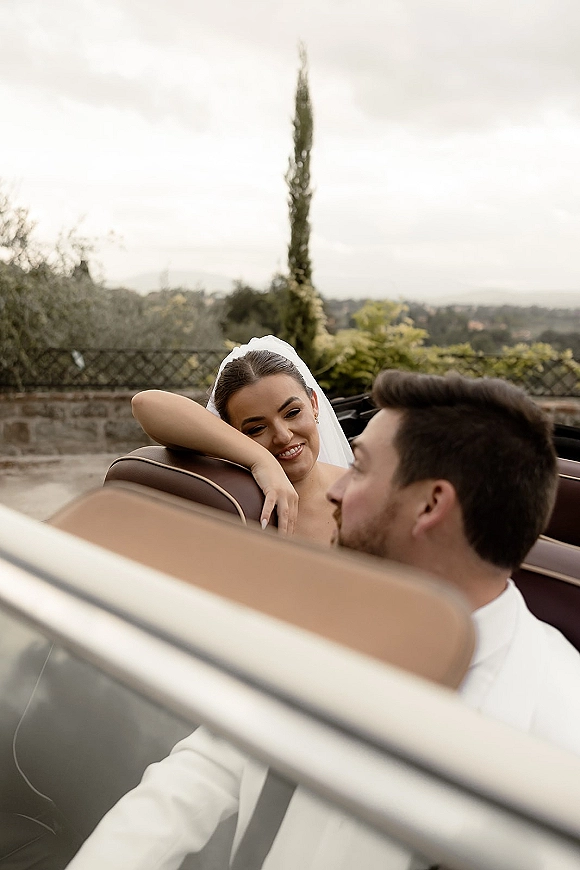 Couple portrait in a wedding getaway car, bride in convertible with veil beside groom driving past a stone wall under overcast sky