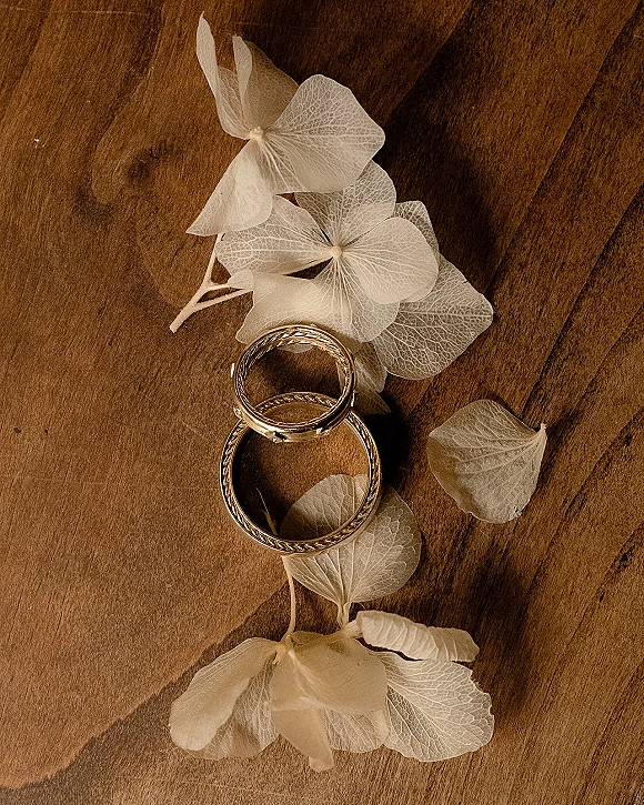 Wedding rings in a wedding ring flat lay, two gold bands resting on a wood tabletop with dried white flowers and scattered petals