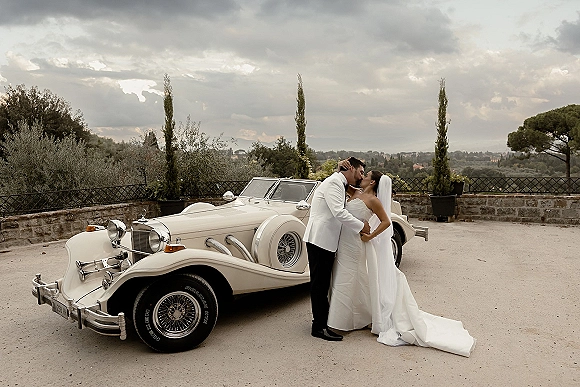 Wedding kiss as bride in strapless dress and veil leans into groom in white tuxedo beside a vintage convertible on a stone terrace