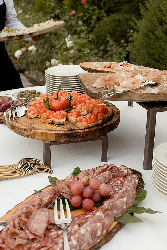 Wedding appetizer table with a wedding charcuterie board featuring prosciutto, salami, bruschetta, grapes, and olives amid garden greenery