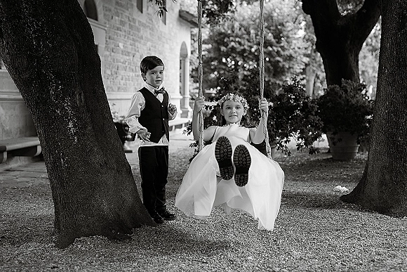 Flower girl and ring bearer pose by a swing, her flower crown and tulle dress beside his vest and bow tie near trees and a brick building
