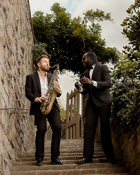 Wedding musicians in tuxedos holding a saxophone on outdoor stone stairs by a rustic wall and wooden gate with greenery behind