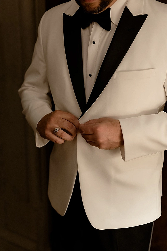 Groom portrait in a white tuxedo jacket with black shawl lapel and bow tie, adjusting his cuff and ring beside an indoor curtain