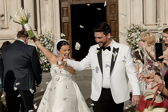 Wedding recessional as bride and groom exit church doors while guests toss rose petals, bride lifting bouquet overhead, veil flowing