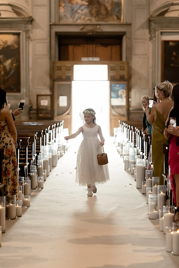Flower girl processional with basket, wearing a white dress and floral crown, walking a candle-lined church aisle runner past guests