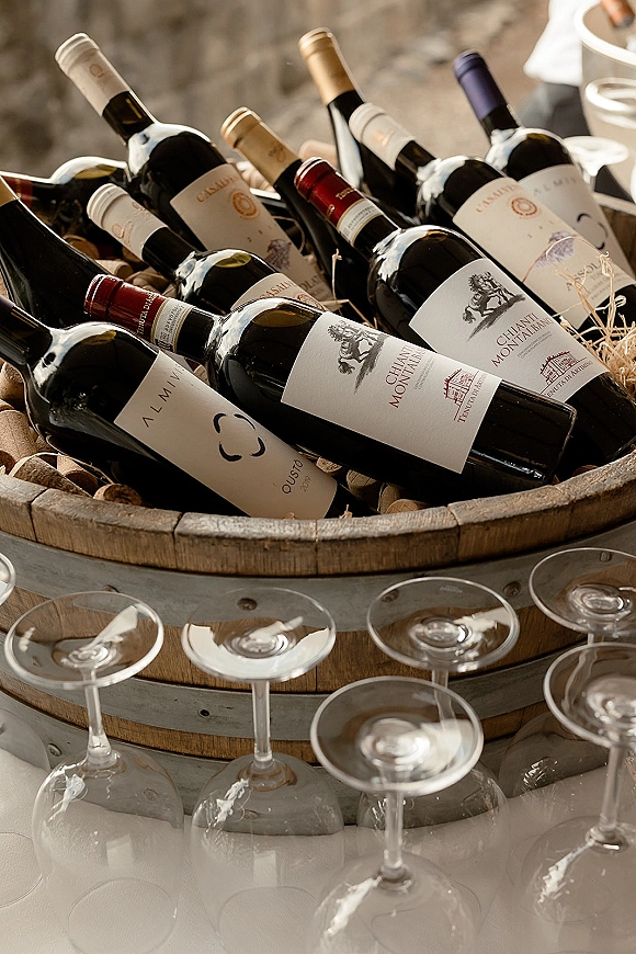 Wine display at a wedding wine station with bottles in a wooden barrel tub, corks and straw filler, and glasses on a tabletop with stone wall backdrop
