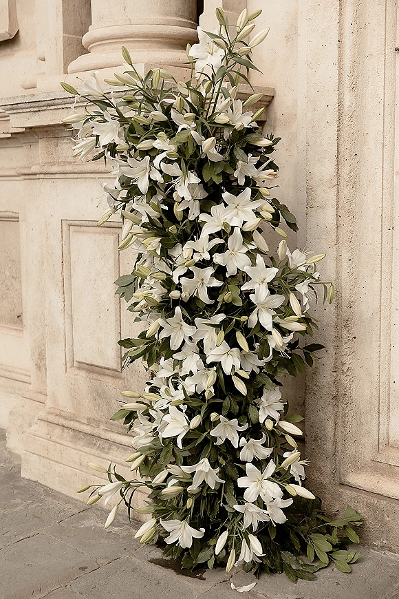 Wedding floral arrangement of white lilies and lush greenery at a stone building entrance with columns on stone pavement