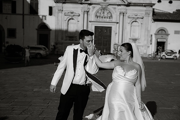 Couple portrait in black and white wedding portrait style, bride in veil and satin dress as groom in white tux kisses her hand by church facade