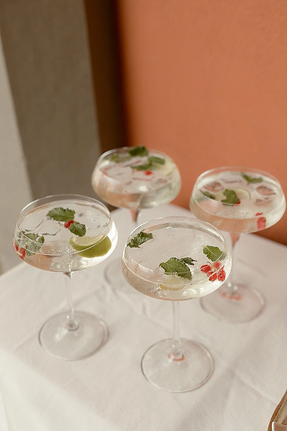 Wedding cocktails in coupe glasses with clear drinks, ice, mint and lime garnishes, plus red fruit accents on a white tablecloth by a peach wall
