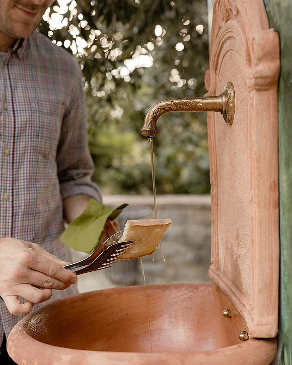 Honey favor station with honey wedding favors in a wooden barrel dispenser with brass spigot, tongs, and cheese beside greenery and stone wall