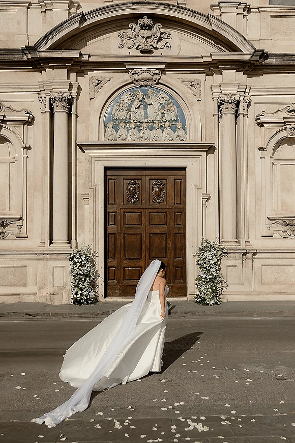 Bridal portrait of a bride with long veil in a strapless wedding dress, back view at stone church doors with petals on ground