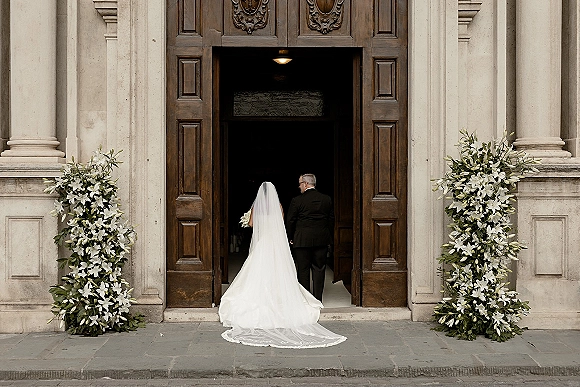 Wedding processional with bride from behind in a long veil and train, holding a bouquet beside a groom in tuxedo at carved church doors