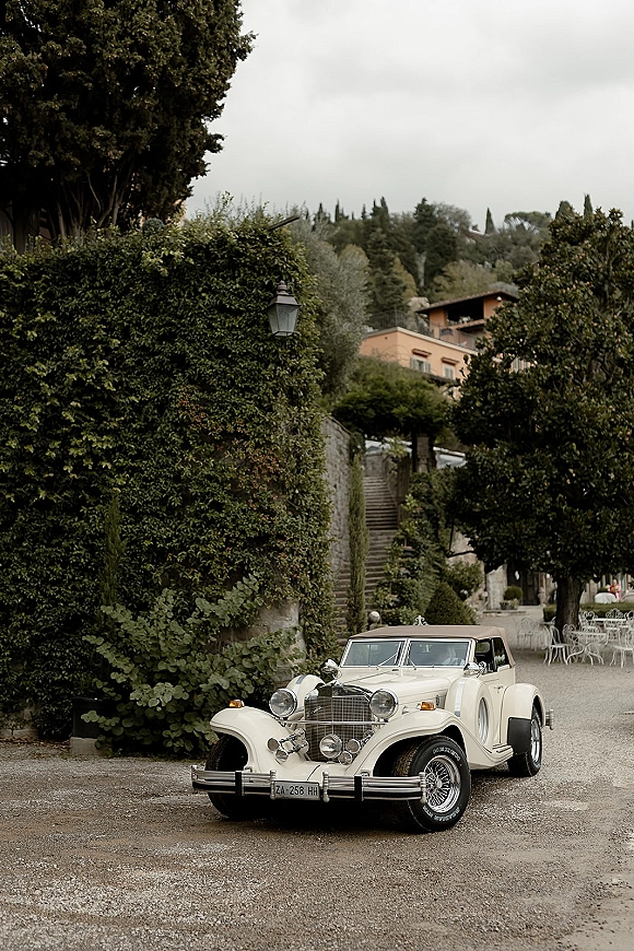 Wedding getaway car, a white vintage convertible with chrome grille and wire wheels parked by an ivy-covered wall and stone steps