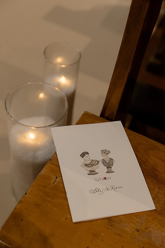 Wedding program displayed on a wooden chair, an illustrated ceremony program card beside glass cylinder candles along a white aisle runner